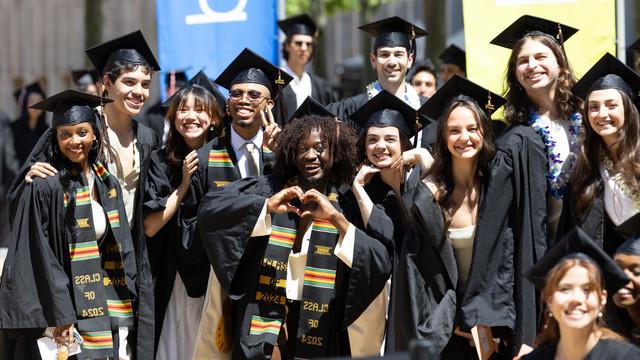 一群茱莉亚学院的毕业生，戴着黑色的帽子，穿着黑色的礼服，一起摆姿势，微笑着拍照. 一些毕业生正在用手做心形图案，而另一些人则戴上花环和五颜六色的披肩. 他们在庆祝毕业，以喜悦的表情和亲密的情谊为标志.